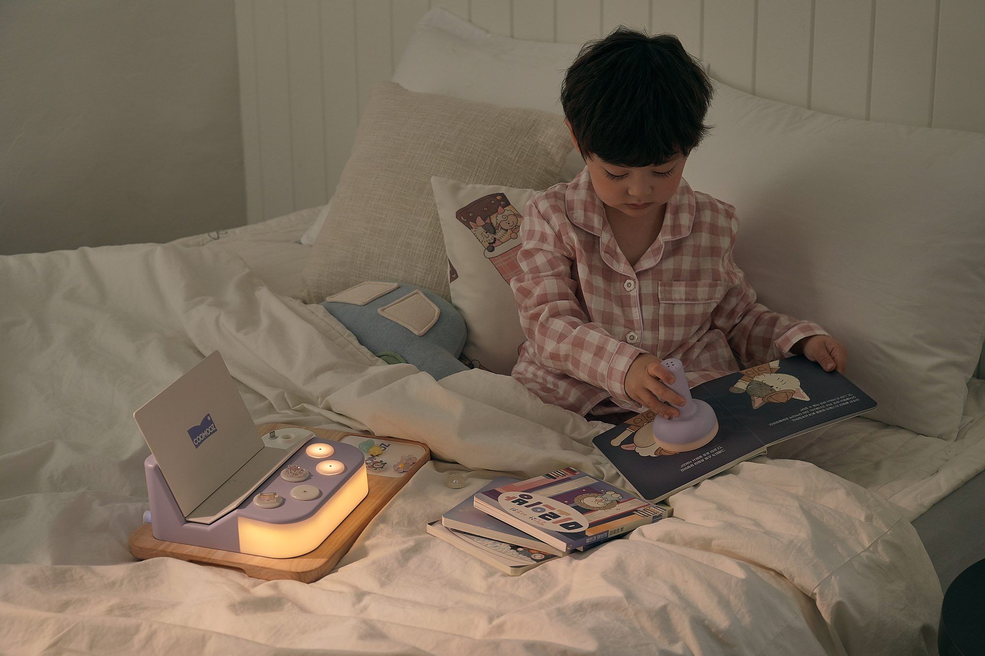 침대 위에서 아이가 COOMOOZ 손전등으로 동화책을 비추고, 옆에 COOMOOZ 스테이션 불빛이 켜져 있는 모습